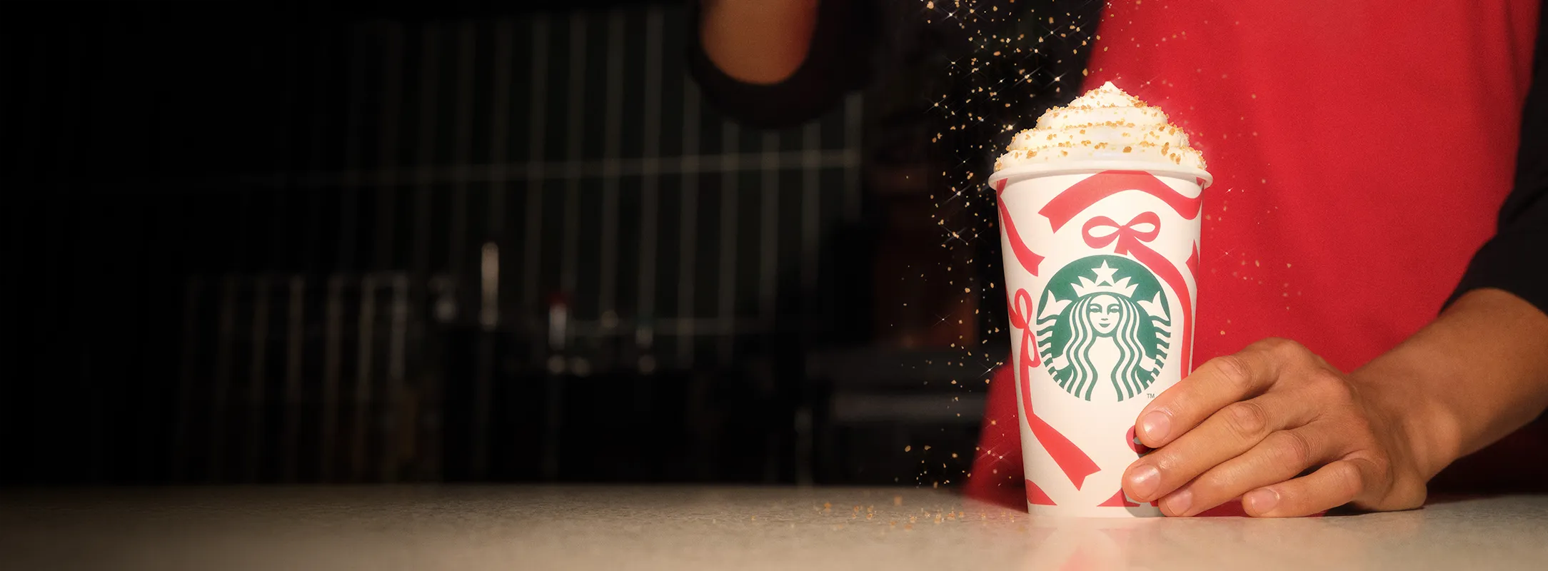 A barista in a red apron holding a Toffee Nut Latte served in a Starbucks Holiday cup. They are serving the drink and shaking a dusting of Toffee Nut flavoured sprinkles on to a layer of whipped cream which tops the drink.