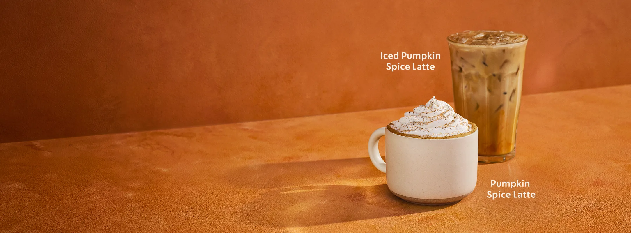 Two Starbucks pumpkin spice drinks on an orange background: a hot Pumpkin Spice Latte in a white mug topped with whipped cream, and an Iced Pumpkin Spice Latte in a clear glass filled with ice.